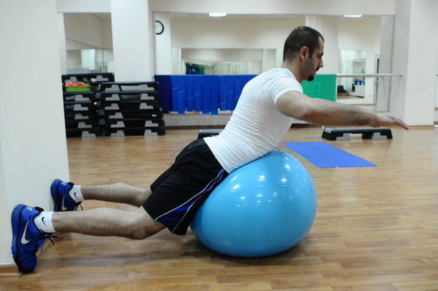 Ayaklarımızı duvara dayayarak yüzüstü pilates topuna uzanıp ellerimizi ileride birleştirip sırtımızı geriye doğru sıkıştırıp dirseklerimizi arkaya doğru alıyoruz.